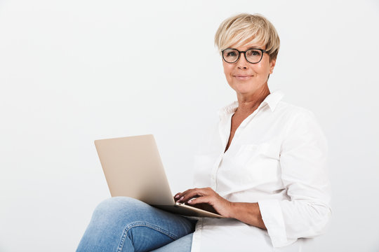 Image of pleased adult woman wearing eyeglasses smiling while sitting with laptop computer
