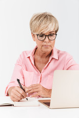 Portrait of mature middle-aged woman with short blond hair wearing eyeglasses using laptop computer and notebook