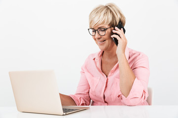 Portrait of caucasian middle-aged woman wearing eyeglasses talking on cellphone and using laptop computer