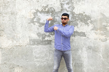 Obraz na płótnie Canvas Portrait of funny handsome bearded young man in checkered blue shirt and sunglasses standing against concrete gray wall. pointing his bicep and showing that his strong.