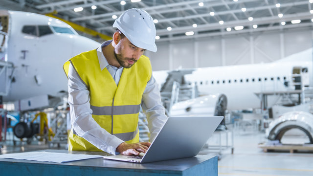 Engineer In Safety Vest Working On Computer In Aircraft Maintenance Hangar