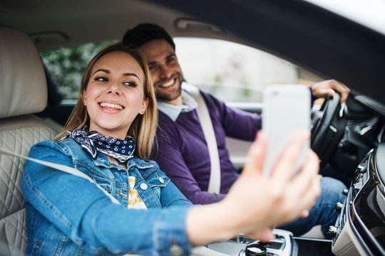 Happy young couple with smartphone sitting in car, taking selfie.
