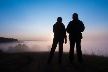 silhouette of two peple looking at sunrise on foggy valley