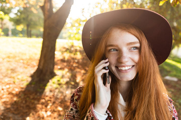 Happy pleased cutie young student redhead girl in autumn park using mobile phone.