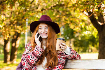 Positive pleased happy cheery young student redhead girl in autumn park talking by mobile phone drinking coffee.
