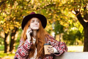 Confused sad young student redhead girl in autumn park talking by mobile phone drinking coffee.