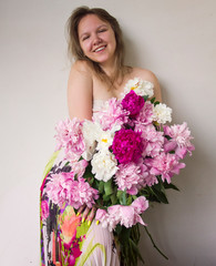 young cute  girl in dress  is standing with  big bouquet of peonies, looking straight and smiling happy on white wall background, romantic female concept, free space