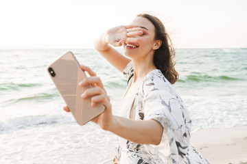 Pleased happy young beautiful woman at the beach walking in a beautiful sunny morning take a selfie by mobile phone.