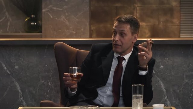 Waist-up shot of smiling middle-aged Caucasian male executive in business suit, white shirt and tie sitting in private lounge holding drink and smoking cigar in his hands and talking with colleagues