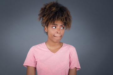 Photo of amazed puzzled young African American female with ginger hair knot, curves lips and has worried look, sees something awful in front, isolated on white background, dressed in jean overalls.