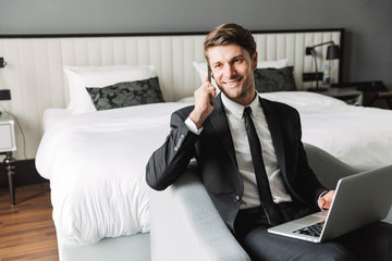 Happy young smiling businessman indoors at home using laptop computer talking by mobile phone.
