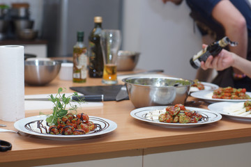 A cook in an apron sets the table for several people, laying out tomatoes in a caprese salad with pesto