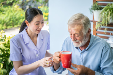 Nurse assist elderly senior man to drink coffee with mug in hand at nursing home