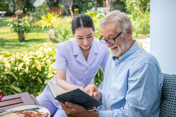 Senior elderly man reading book with nurse during breakfast in garden at nursing home