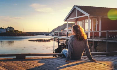 Rolgordijnen Beautiful young  girl traveler sitting on wooden pier on the background of traditional red rorbu traveling to Lofoten Norway © olezzo