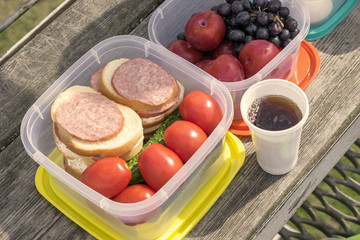 On a wooden table in a plastic container are vegetables: tomatoes and cucumbers, as well as sandwiches with sausage. In the container are fruits: plums and grapes. Next to the food are cups of tea.	