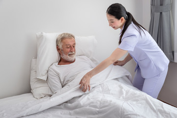 Fototapeta premium Happy nurse cover elderly man with blanket in bedroom at nursing home