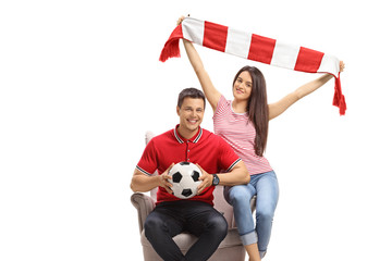 Young male and female soccer fans with a football and a scarf sitting in an armchair