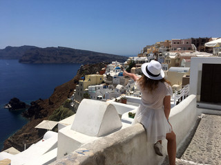 A young woman in a white dress and a white hat is looking at the sea. A tourist woman on vacation in Oia