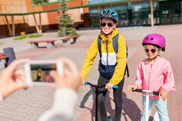 Naklejka premium education, childhood and people concept - happy school children in helmets with backpacks and scooters being photographed by smartphone outdoors