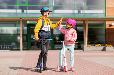 education, childhood and people concept - happy school children in helmets with backpacks riding scooters and making high five outdoors