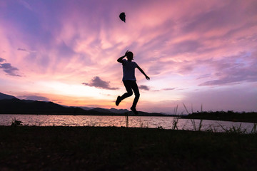 Silhouette of a happy lady jumping and throwing a hat in the air while dramatic sunset sky with beautiful clouds. Traveling and vacation concept.