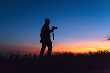 Silhouette of man taking pictures with his camera while sunset hour; against twilight sky over a mountain. Traveling and vacation concept.