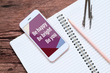 Smartphone mockup for your business quotes with Be happy Be bright Be you text on screen and open notebook on the grunge wood desk table .