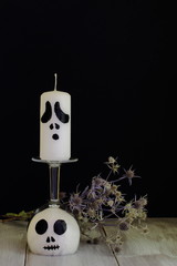 Goblet stylized as charming ghost, white wax candle in shape of Halloween character and thistle on natural wooden surface against black wall. Concept - Нalloween, autumn, holiday, symbol, tradition.