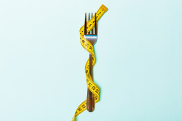Close up of fork wrapped with tape measure on blue background. Top view of obesity concept - obrazy, fototapety, plakaty
