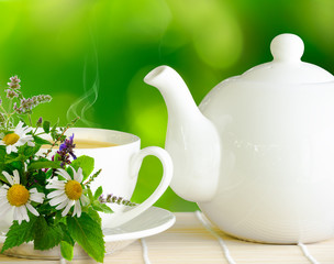 White teapot and cup of herbal tee with bunch of chamomile, mint and other herbs on green nature blurred background