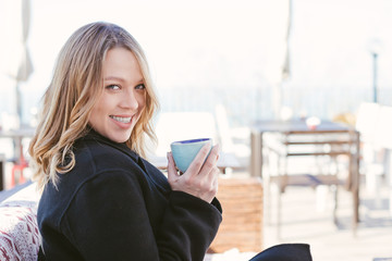 Portrait of curvy smiling blonde woman holding blue coffee mug sitting outdoors at cafe terrace, wearing black coat