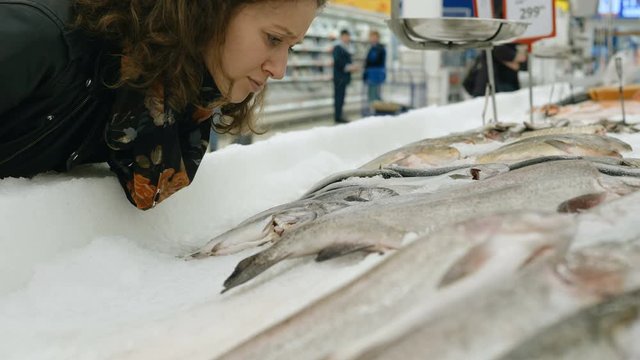 curly girl sniffs fresh fish lying on ice and winces from the smell in a supermarket