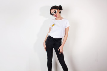 Fototapeta premium young funny brunette girl in a jeans and white shirt with sunglasses is standing and showing tongue on a white wall background, free space, fashion concept