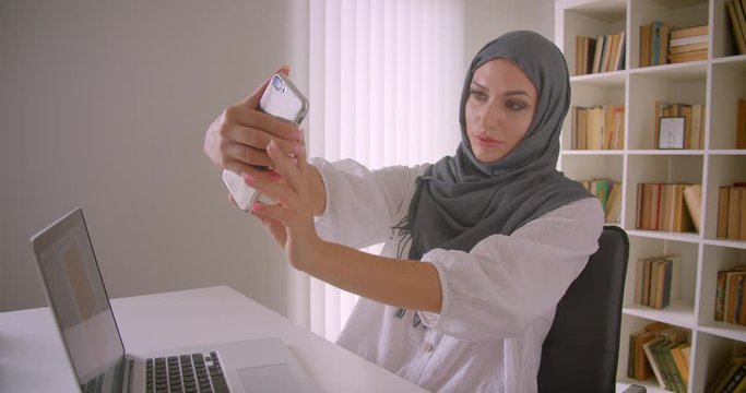 Closeup portrait of young attractive muslim businesswoman in hijab taking selfies on phone posing sitting in front of laptop in office indoors