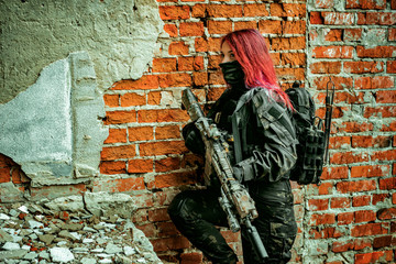 Red-hair airsoft woman in uniform with machine-gun, stand beside brick wall inside broken building