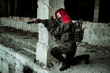 Red-hair airsoft woman in uniform with machine gun on one knee beside column. Soldier aims at the sight stand inside of broken building