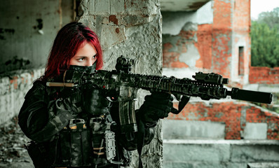 Fototapeta premium Airsoft red-head woman in uniform with machine gun standing on balkony. Soldier aim at the sight