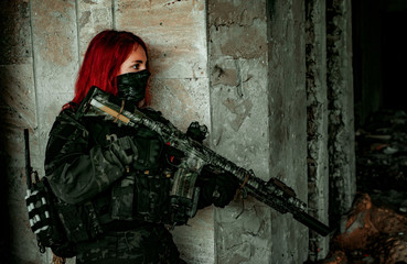 Fototapeta premium Airsoft red-hair woman in uniform with machine gun stand beside concrete wall. Soldier lurk inside broken building. Side view