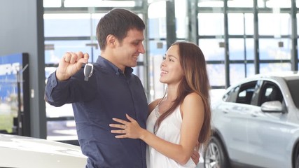 Happy caucasian couple hugging in front of new car, holding the keys, at the car dealership. Car Purchase and Service Concept.