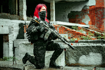 Airsoft red-hair woman in uniform with machine gun standing on knee. Soldier on ruine. Side view