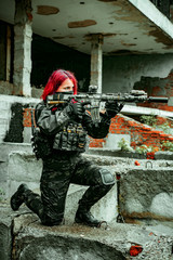 Airsoft red-hair woman in uniform with machine gun standing on knee. Soldier aims at the sight on ruine. Vertical photo side view