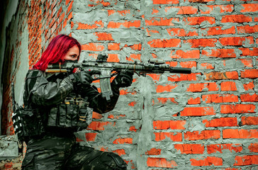 Airsoft red-hair woman in uniform with machine gun beside brick wall. Close up soldier aims at the sight on the ruins. Horizontal photo side view