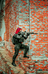 Fototapeta premium Airsoft red-hair woman in uniform with machine gun beside brick wall. Soldier aims at the sight on the ruins. Vertical photo side view