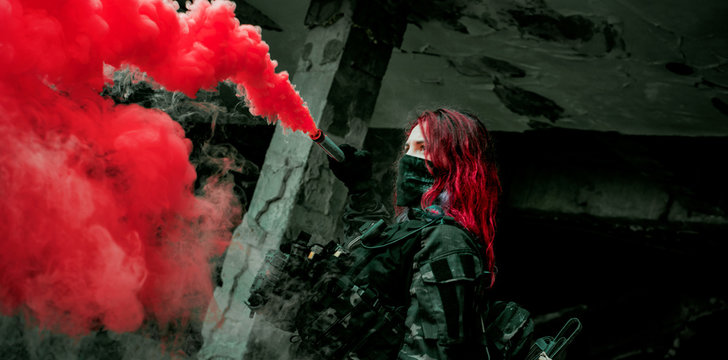 Airsoft red-hair woman in uniform with red smoke in hand. Close up soldier in broken building. Horizontal photo