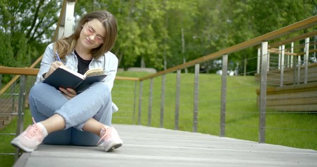 close-up of dark-haired young girl with glasses, sitting in the Park on the bridge cross-legged, she reads a book and something in it marks