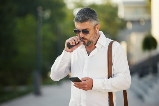 Middle-aged bearded thinking serious concentrated business man standing and smoking electronic cigarette. Blows smoke through the mouth. In-ear wireless earphone. Office centre background 