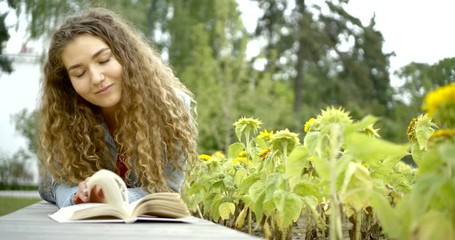close-up portrait of a curly girl who lies in the garden against the background of sunflowers, reading a book and smiling.