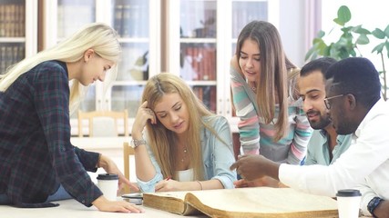 Mixed-race group of university students doing linguistic researches, turning over the pages of big old reference textbook at library.
