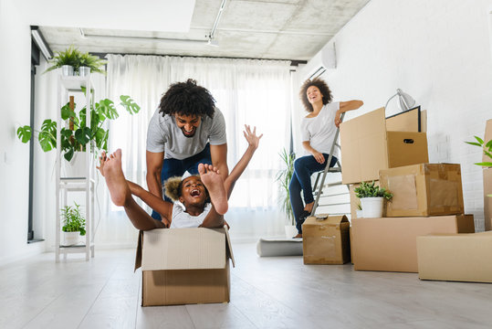 Happy playful  African American family moving in new apartment, little preschooler daughter sitting in cardboard boxes, father rolling her,  purchase property concept